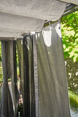 Graues Vorhang-Set im Freien, aufgehängt an einer Pergola, mit Blick auf die Seite und umgeben von grünen Pflanzen.