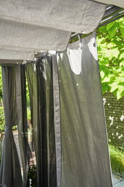 Graues Vorhang-Set im Freien, aufgehängt an einer Pergola, mit Blick auf die Seite und umgeben von grünen Pflanzen.