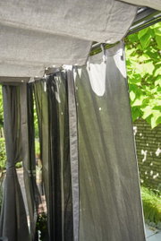 Graues Vorhang-Set im Freien, aufgehängt an einer Pergola, mit Blick auf die Seite und umgeben von grünen Pflanzen.
