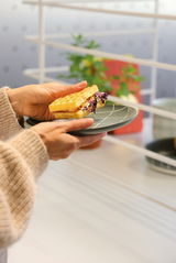 Eine Person hält einen flachen Teller mit einem Waffelsandwich in der Hand, seitliche Perspektive.