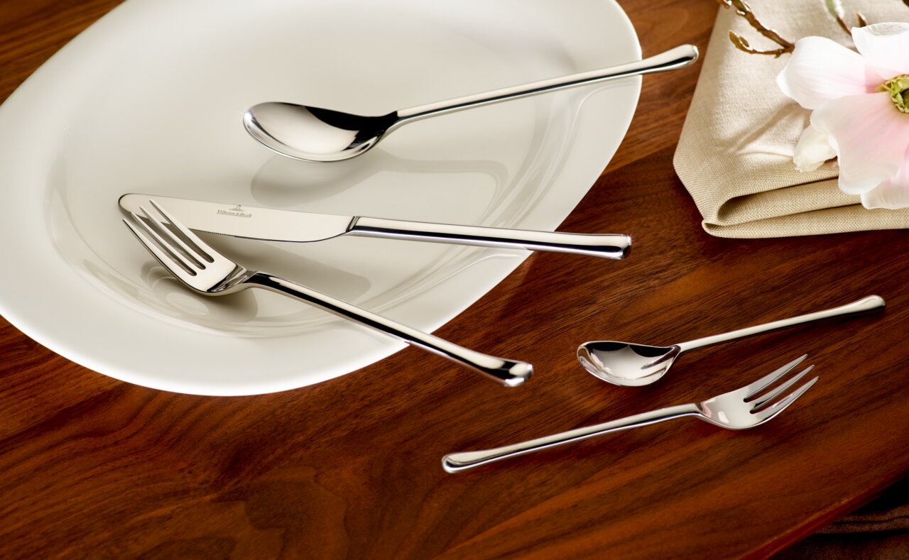 Elegantes 30-teiliges Besteckset aus Edelstahl, auf einem weißen Teller und Holztisch arrangiert, mit einer Blume und Serviette im Hintergrund, aus der Vogelperspektive fotografiert.