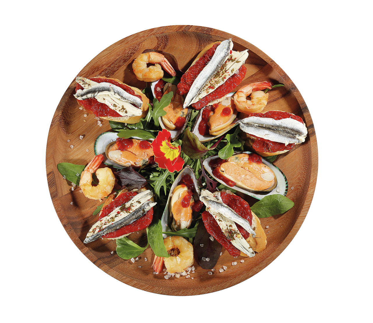 Vogelperspektive auf einen runden Snackteller aus Akazienholz, dekoriert mit Meeresfrüchten, Gemüse und essbaren Blüten.