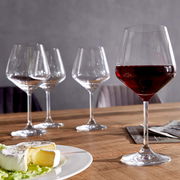 Vier Burgundergläser auf einem Holztisch, eines gefüllt mit Rotwein, im Vordergrund ein Teller mit Käse. Perspektive: seitlich.