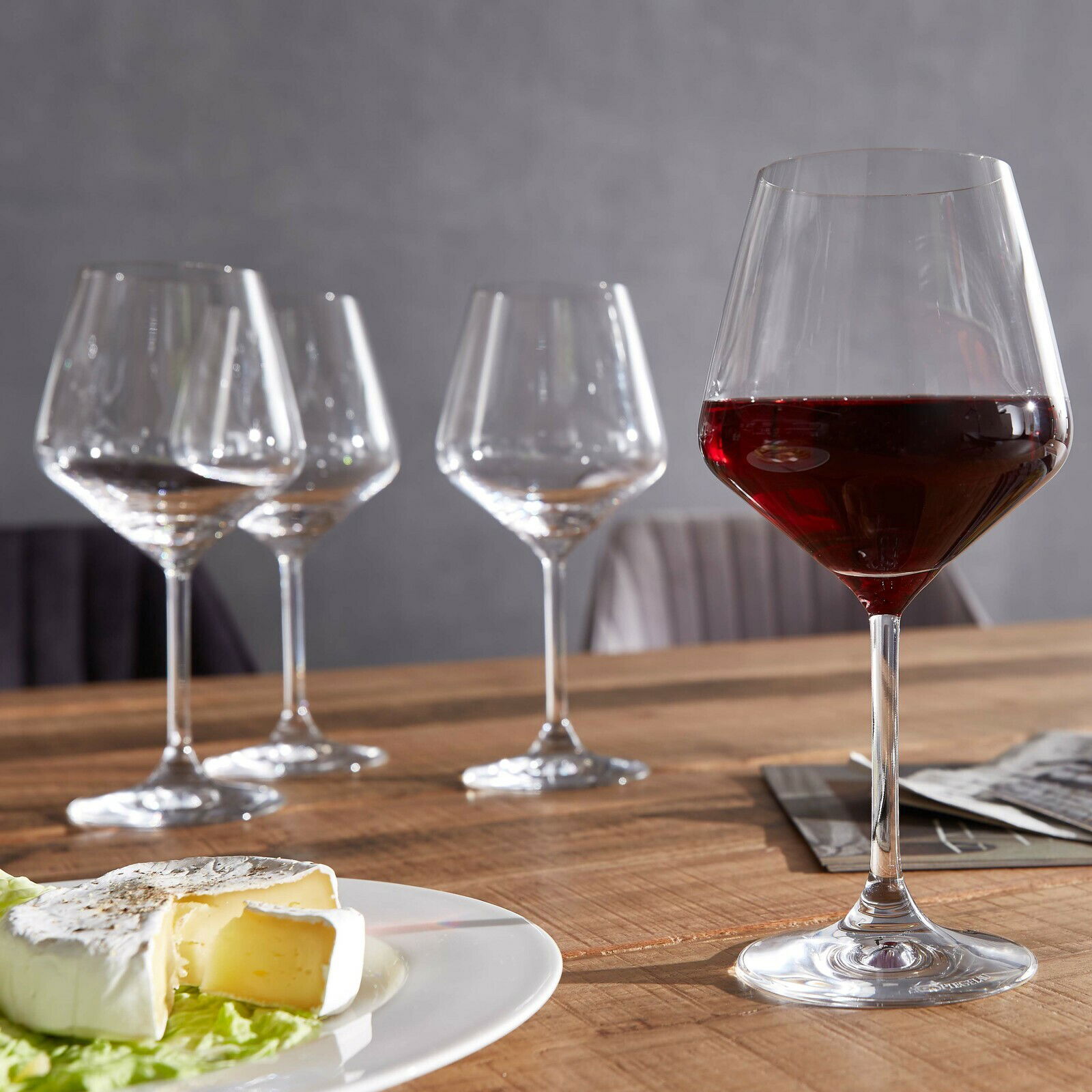 Vier Burgundergläser auf einem Holztisch, eines gefüllt mit Rotwein, im Vordergrund ein Teller mit Käse. Perspektive: seitlich.