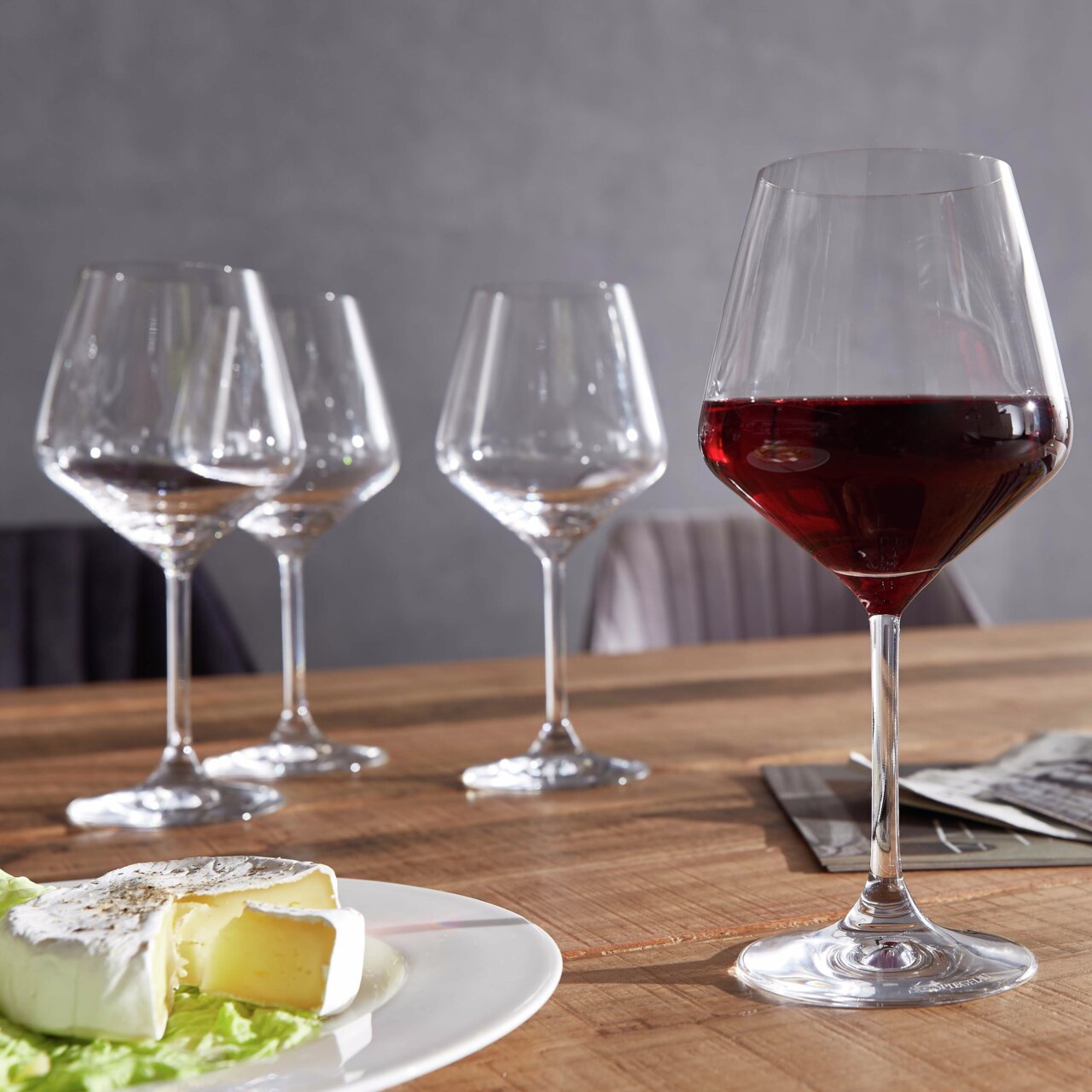 Vier Burgundergläser auf einem Holztisch, eines gefüllt mit Rotwein, im Vordergrund ein Teller mit Käse. Perspektive: seitlich.