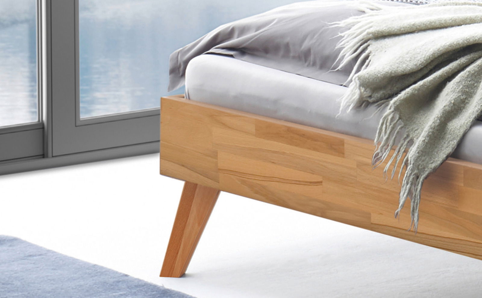 Nahaufnahme eines modernen Bettgestells aus Holz mit schrägen Beinen, von der Seite betrachtet. Das Bett ist mit einer grauen Decke dekoriert und steht vor einem großen Fenster.