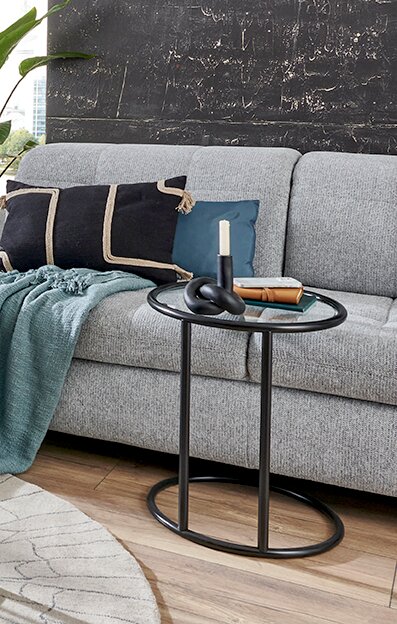 Moderner schwarzer Beistelltisch mit runder Glasplatte vor grauem Sofa mit Kissen und Decke, seitliche Perspektive im Wohnzimmer