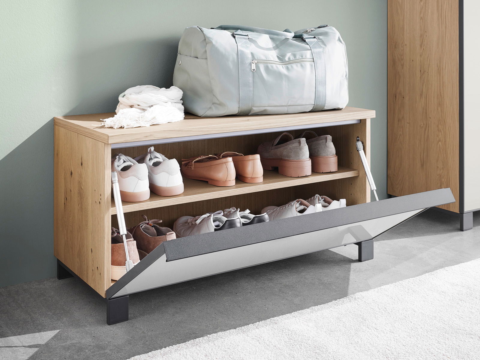 Garderobenbank aus Holz mit geöffneter Klappe, die Schuhe auf zwei Regalböden zeigt, mit einer Tasche und einem Schal darauf, seitliche Perspektive.