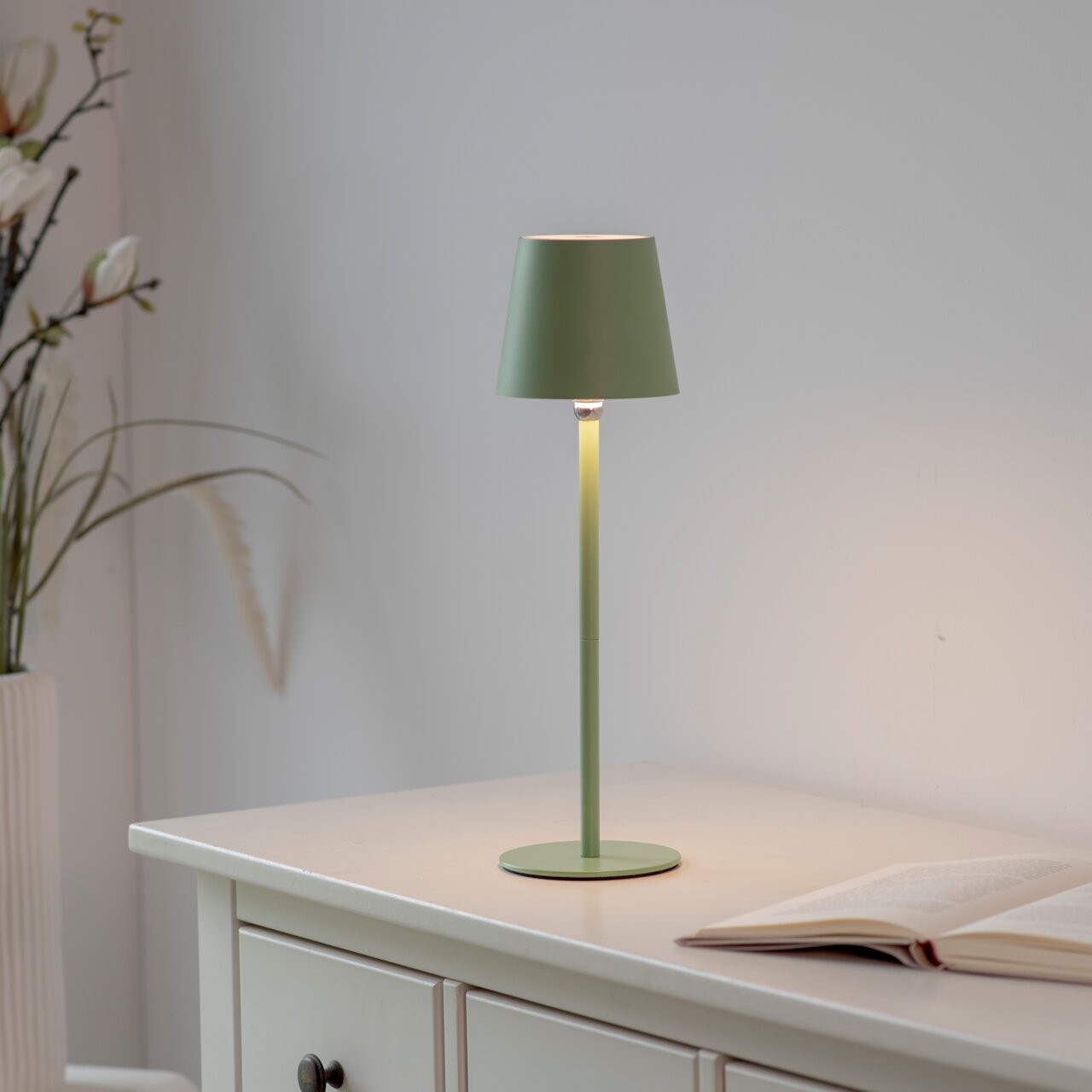 Grüne Akku-Tischleuchte BOTERIA auf einem weißen Tisch, seitliche Perspektive, mit einer offenen Blume im Hintergrund.