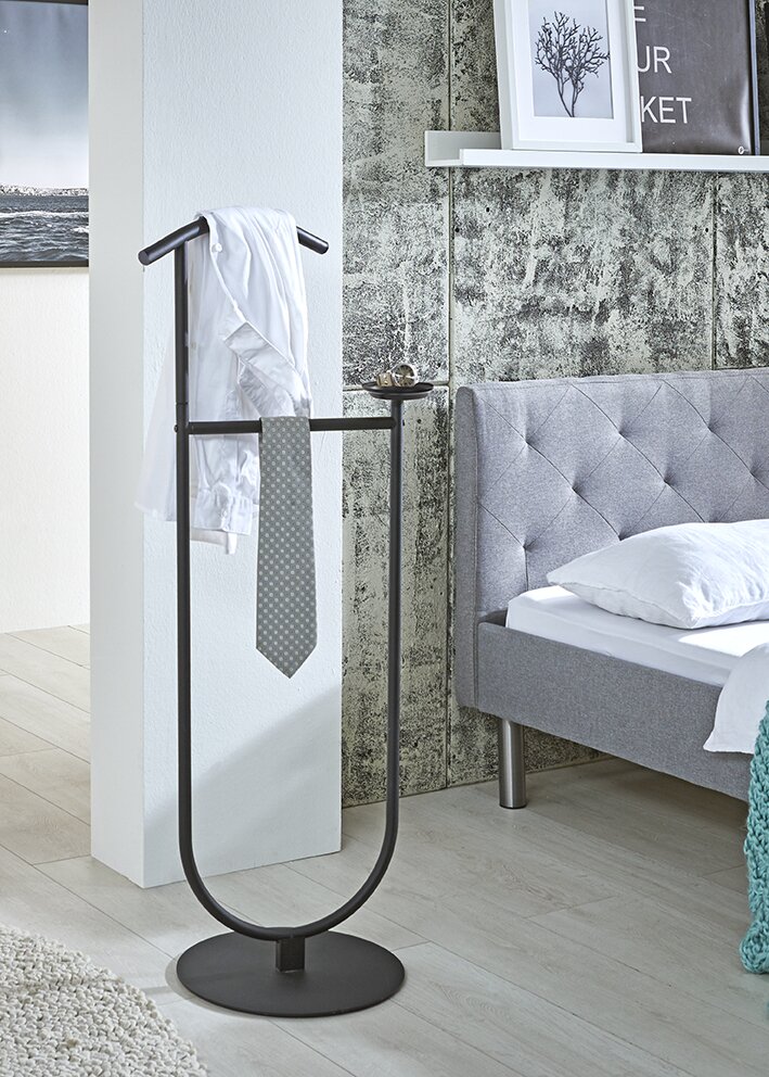 Zurbrüggen Herrendiener Schwarzer, freistehender Kleiderständer/Valet-Stand mit rundem Standfuß und integrierter Ablageschale, mit Hemd und Krawatte dekoriert, im Schlafzimmer neben einem grauen Bett; perspektivisch in leicht schräger Vorderansicht.