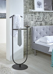 Schwarzer, freistehender Kleiderständer/Valet-Stand mit rundem Standfuß und integrierter Ablageschale, mit Hemd und Krawatte dekoriert, im Schlafzimmer neben einem grauen Bett; perspektivisch in leicht schräger Vorderansicht.