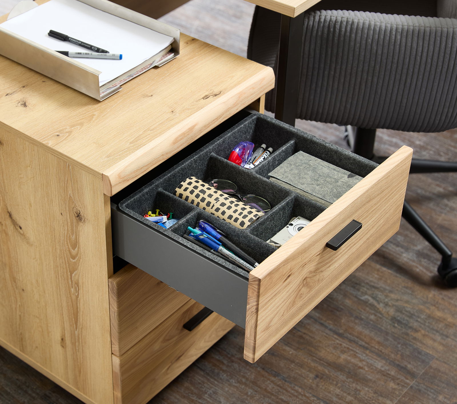 Rollcontainer aus Holz mit geöffneter Schublade, gefüllt mit Büroartikeln, aus der Perspektive von schräg oben.