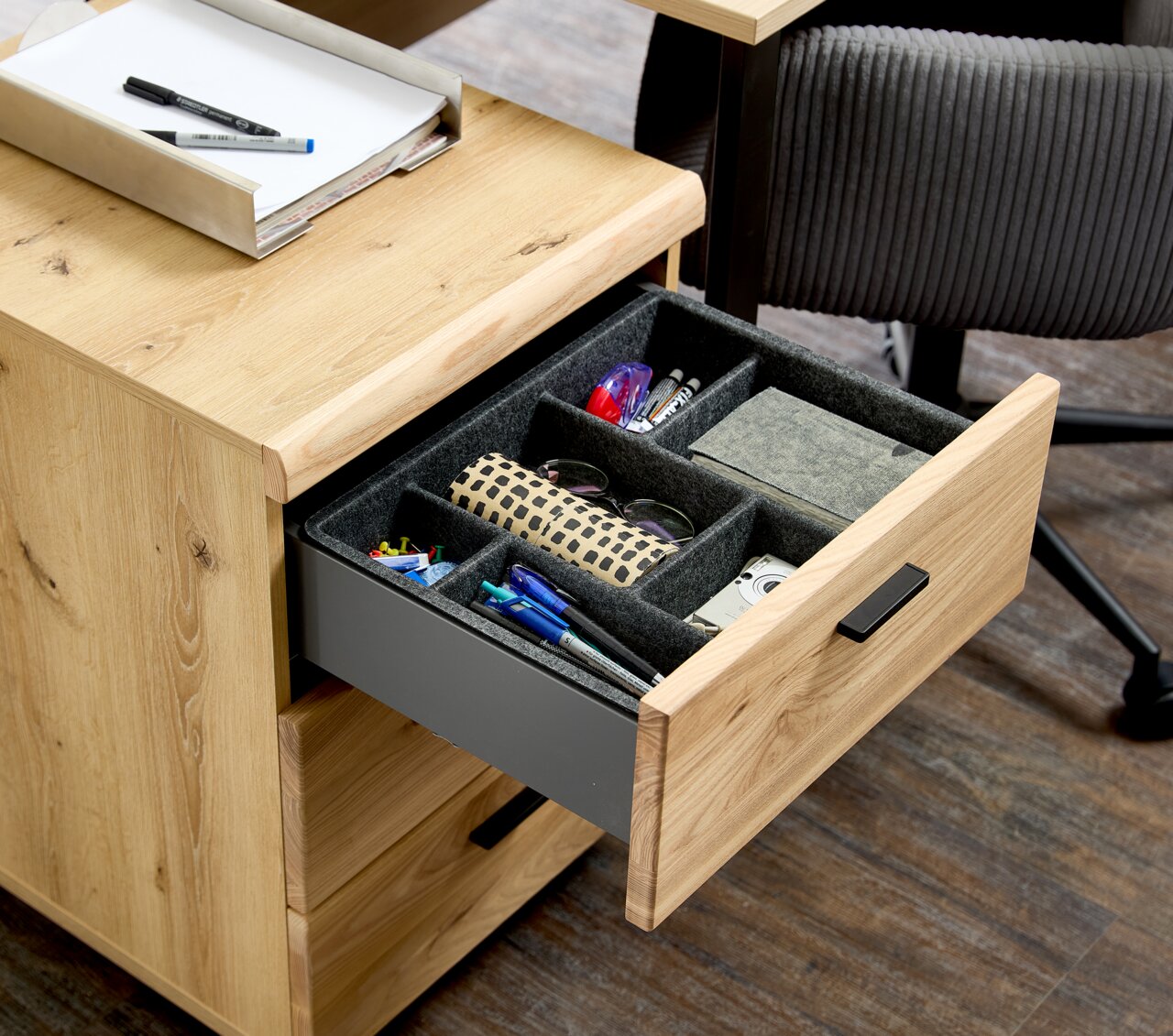 Rollcontainer aus Holz mit geöffneter Schublade, gefüllt mit Büroartikeln, aus der Perspektive von schräg oben.