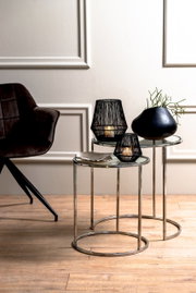 Twee ronde bijzettafels met glazen bladen en metalen frames, versierd met zwarte kandelaars en een vaas, gezien vanuit de zijkant.