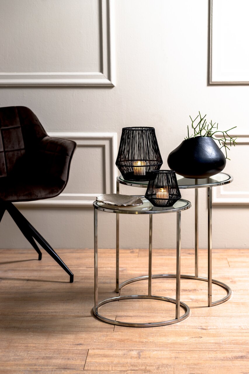 Twee ronde bijzettafels met glazen bladen en metalen frames, versierd met zwarte kandelaars en een vaas, gezien vanuit de zijkant.