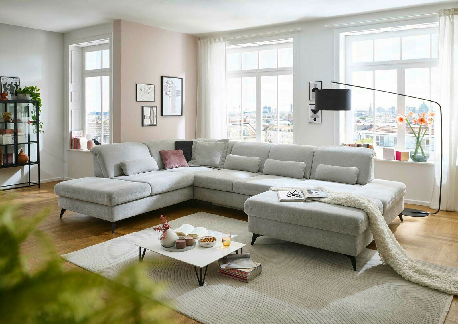 Große, moderne Polsterrundecke in einem hellen Wohnzimmer mit großen Fenstern, die viel Tageslicht hereinlassen. Die Couch ist in einem hellgrauen Stoff bezogen und bietet viel Platz zum Entspannen. Perspektive von vorne links.