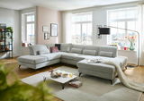 Große, moderne Polsterrundecke in einem hellen Wohnzimmer mit großen Fenstern, die viel Tageslicht hereinlassen. Die Couch ist in einem hellgrauen Stoff bezogen und bietet viel Platz zum Entspannen. Perspektive von vorne links.