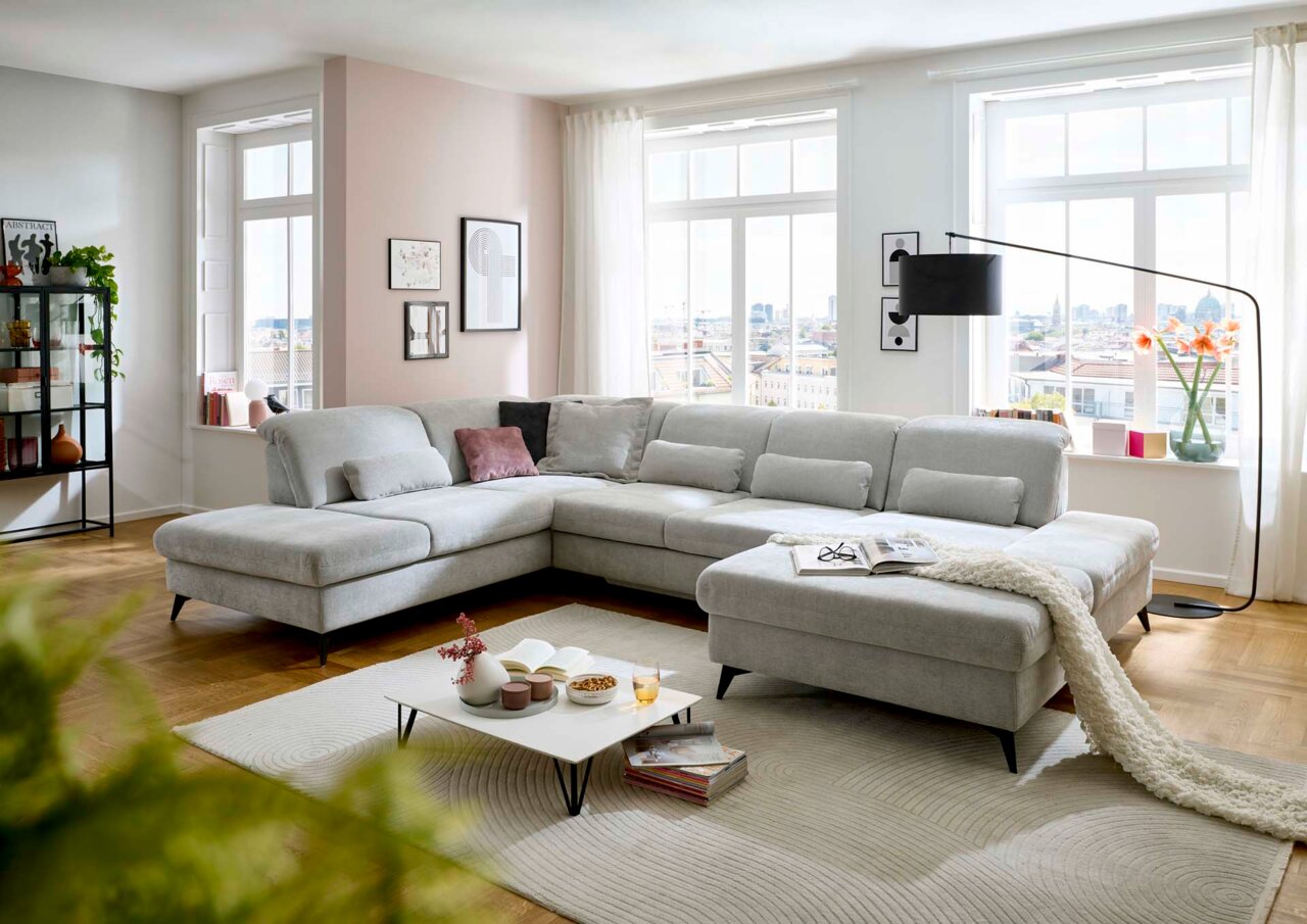 Beldomo Style Wohnlandschaft BREMEN Große, moderne Polsterrundecke in einem hellen Wohnzimmer mit großen Fenstern, die viel Tageslicht hereinlassen. Die Couch ist in einem hellgrauen Stoff bezogen und bietet viel Platz zum Entspannen. Perspektive von vorne links.