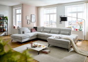 Große, moderne Polsterrundecke in einem hellen Wohnzimmer mit großen Fenstern, die viel Tageslicht hereinlassen. Die Couch ist in einem hellgrauen Stoff bezogen und bietet viel Platz zum Entspannen. Perspektive von vorne links.