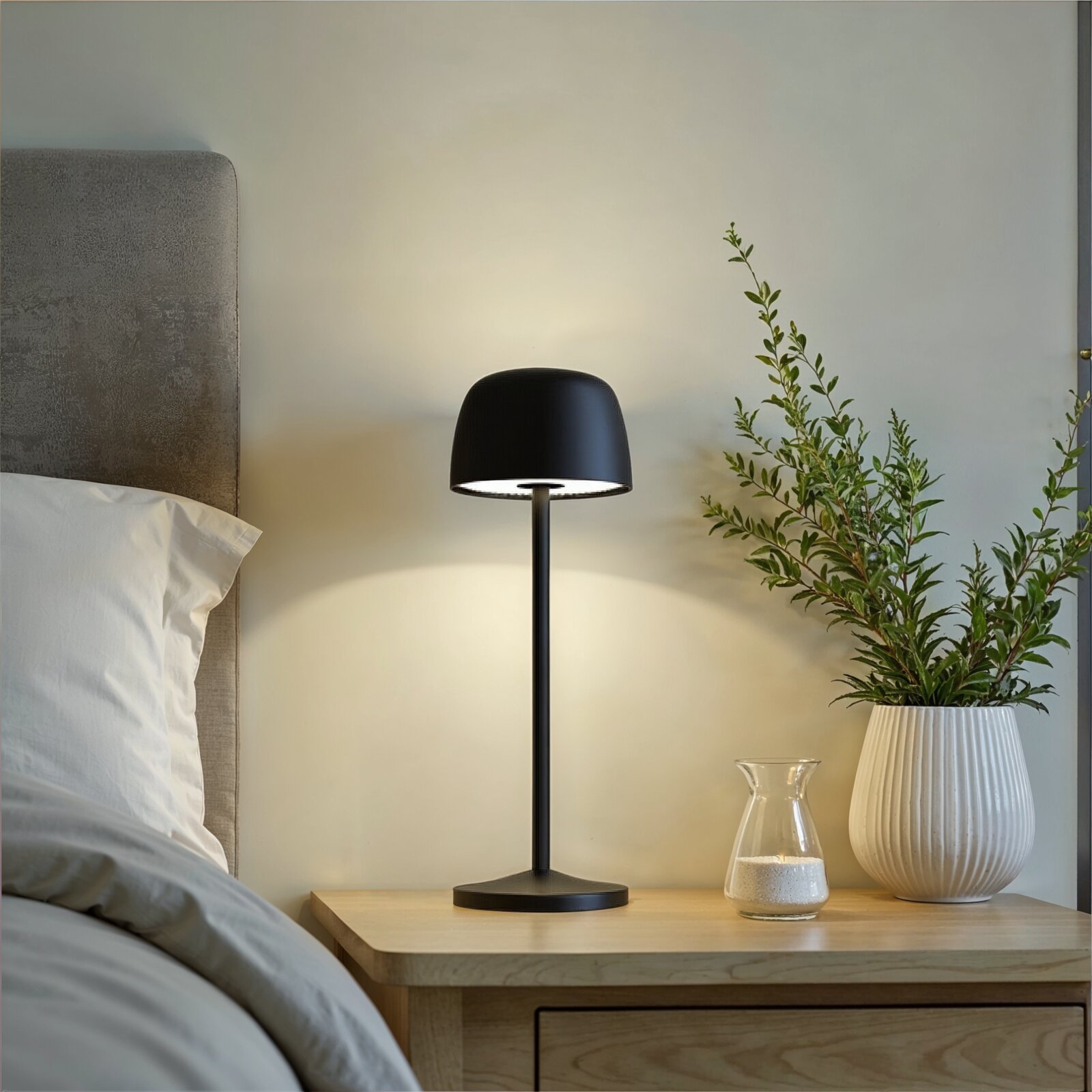 Schwarze, moderne LED-Tischlampe mit kuppelförmigem Schirm auf einem Holz-Nachttisch neben Bett und Vase, frontal aus leichter Seitenperspektive fotografiert.