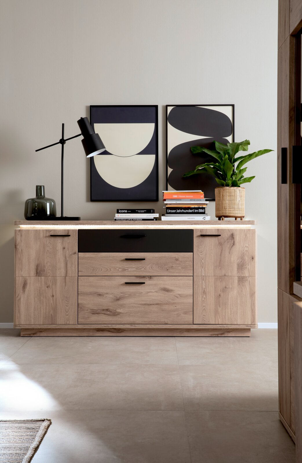 Zurbrüggen Sideboard MODENA Frontale Ansicht einer modernen Holz-Kommode/Sideboard in Eiche-Optik mit schwarzer Schubladenfront und schwarzen Griffen, dekoriert mit Tischlampe, Büchern, Vase und Zimmerpflanze vor abstrakten Wandbildern.