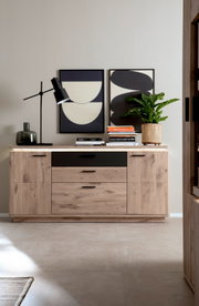 Vooraanzicht van een moderne houten ladekast/dressoir in eikenlook met zwarte ladefronten en zwarte handgrepen, versierd met een tafellamp, boeken, een vaas en een kamerplant tegen een achtergrond van abstracte wandschilderijen.