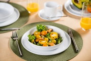 Schräg von oben fotografierter gedeckter Tisch mit grünem Platzset, weißem Teller und Schüssel mit frischem Salat aus grünen Blättern, gelben Tomaten und Kräutern; daneben Besteck sowie Tasse und Gläser mit Orangensaft im Hintergrund.