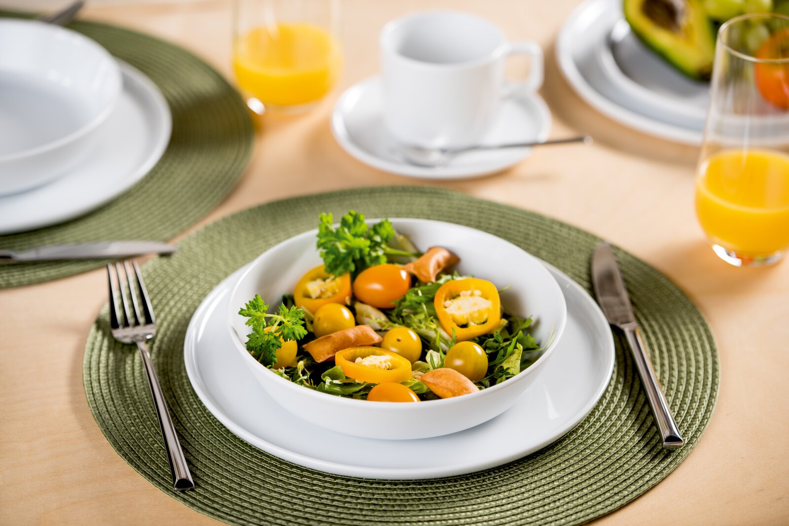 Schräg von oben fotografierter gedeckter Tisch mit grünem Platzset, weißem Teller und Schüssel mit frischem Salat aus grünen Blättern, gelben Tomaten und Kräutern; daneben Besteck sowie Tasse und Gläser mit Orangensaft im Hintergrund.