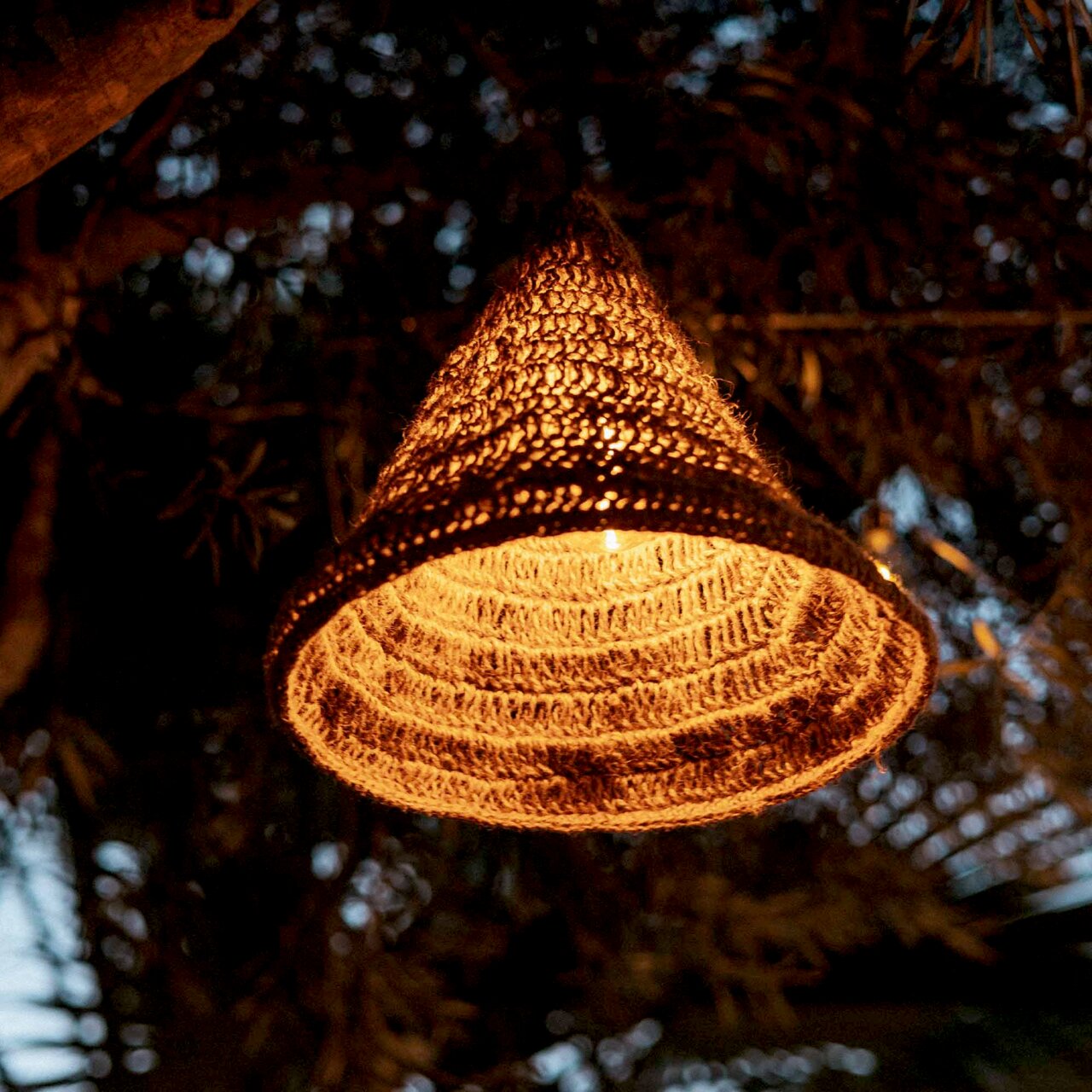 Outdoor-Hängeleuchte aus geflochtenem Material, beleuchtet in einer abendlichen Gartenumgebung, von unten betrachtet.