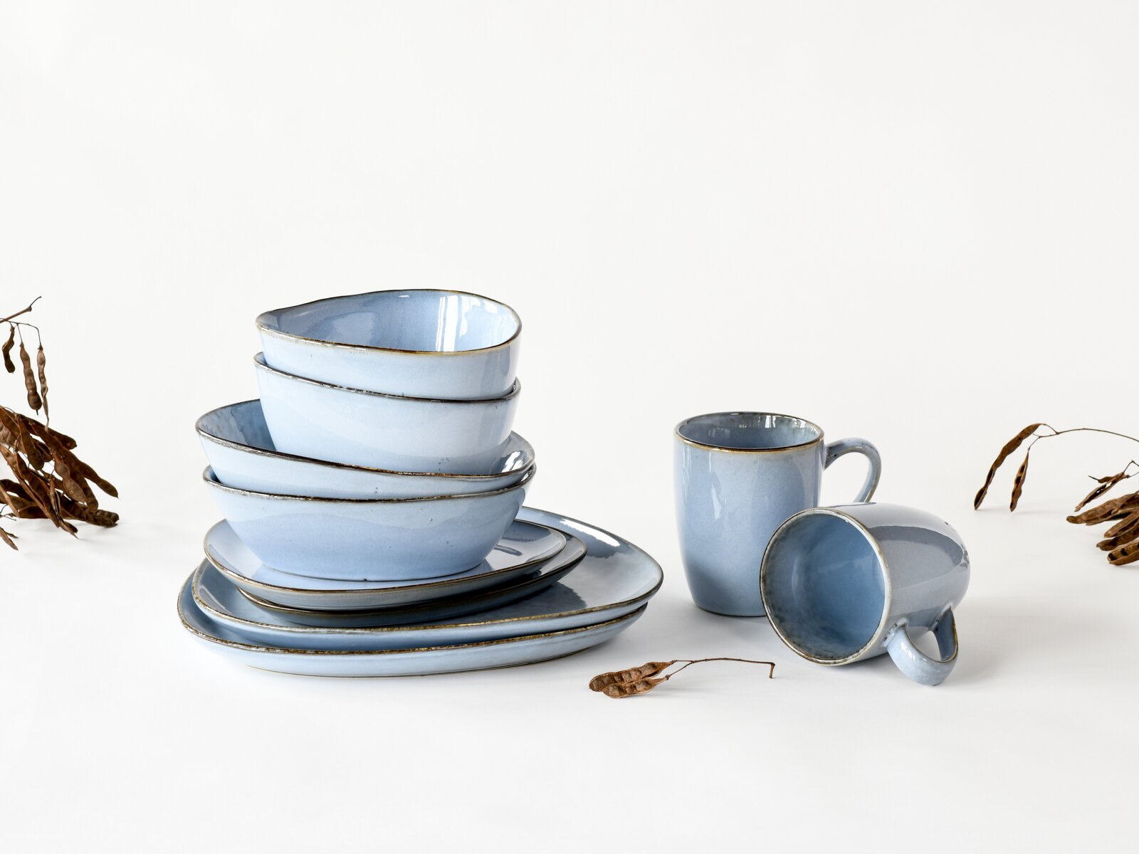 Hellblaues glasiertes Keramik-Geschirrset mit gestapelten Schalen und Tellern sowie zwei Bechern auf weißem Hintergrund, fotografiert in leichter Seitenansicht auf Augenhöhe.