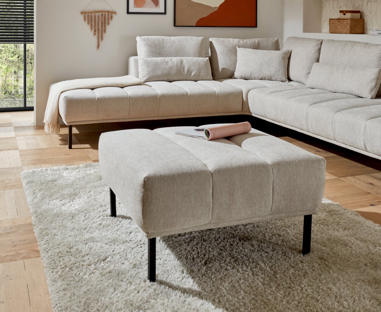 Beiger Hocker mit schwarzem Metallgestell, im Vordergrund eines modernen Wohnzimmers mit Ecksofa, aus der Perspektive von vorne fotografiert.