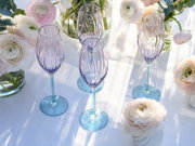 Drei elegante Champagnergläser aus lilafarbenem Glas mit strukturierter Oberfläche und hellblauem Stiel auf weißer Tischdecke, umgeben von zarten Ranunkeln und Eukalyptus; Perspektive: leicht von oben, schräger Blickwinkel.