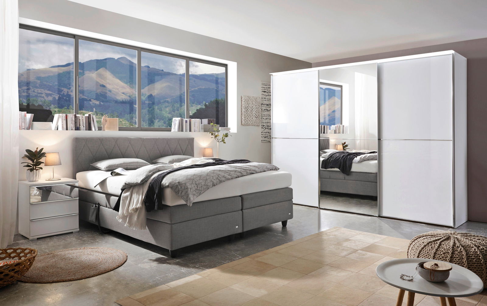 Modernes Schlafzimmer mit einem grauen Boxspringbett, seitlicher Perspektive, neben einem weißen Nachttisch und einem großen weißen Kleiderschrank mit Spiegel.