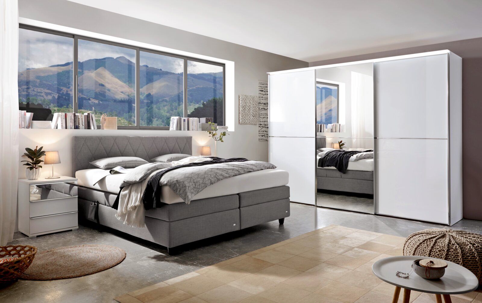 Modernes Schlafzimmer mit einem grauen Boxspringbett, seitlicher Perspektive, neben einem weißen Nachttisch und einem großen weißen Kleiderschrank mit Spiegel.