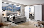 Modernes Schlafzimmer mit einem grauen Boxspringbett, seitlicher Perspektive, neben einem weißen Nachttisch und einem großen weißen Kleiderschrank mit Spiegel.