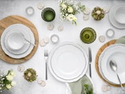 Draufsicht auf eine elegant gedeckte Tafel mit weißem Porzellan-Geschirr (Teller, Schalen und Tasse), grünem Strukturglas, Besteck, Holzuntersetzern, Teelichtern und weißen Blumen auf hellem Hintergrund.