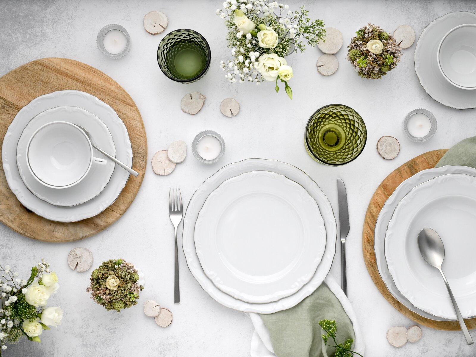 Draufsicht auf eine elegant gedeckte Tafel mit weißem Porzellan-Geschirr (Teller, Schalen und Tasse), grünem Strukturglas, Besteck, Holzuntersetzern, Teelichtern und weißen Blumen auf hellem Hintergrund.