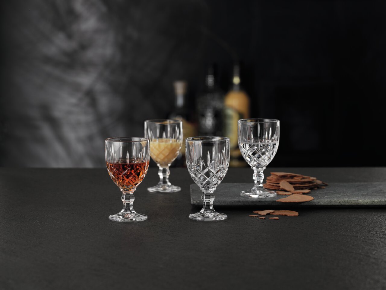 Vier elegante Likörkelche aus Kristallglas, auf einem dunklen Tisch arrangiert, mit einem unscharfen Hintergrund. Zwei Gläser sind gefüllt, zwei leer. Perspektive von vorne.