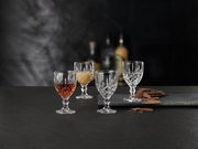 Vier elegante Likörkelche aus Kristallglas, auf einem dunklen Tisch arrangiert, mit einem unscharfen Hintergrund. Zwei Gläser sind gefüllt, zwei leer. Perspektive von vorne.