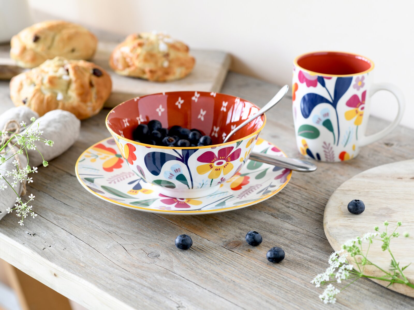 Bunte Keramik-Schüssel mit Blumenmuster auf passendem Teller, gefüllt mit Blaubeeren und Löffel, daneben passende Tasse auf Holztisch – Ansicht leicht von oben in schräger Perspektive
