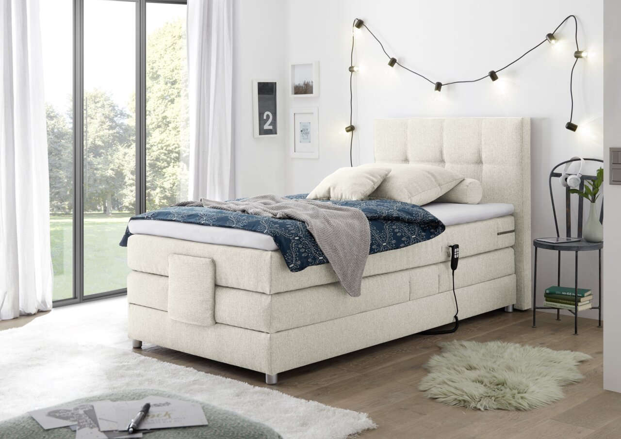 Boxspringbed in een moderne slaapkamer, gezien vanaf de zijkant, met een beige stoffen bekleding, een blauw dekbed en kussens, versierd met een lichtslinger aan de muur.
