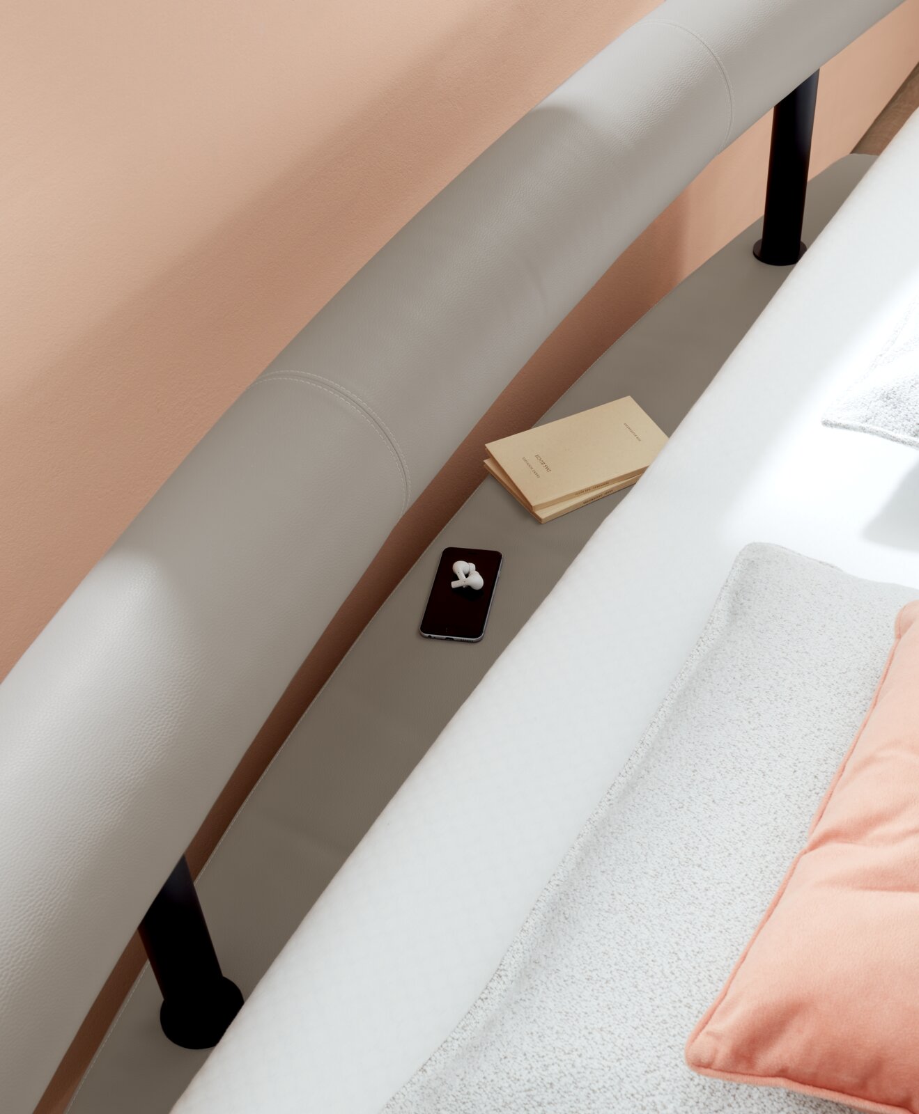 Detailfoto van een gestoffeerd bed met een gebogen hoofdeinde, met daarop een smartphone, een koptelefoon en boeken, gezien vanuit vogelperspectief.