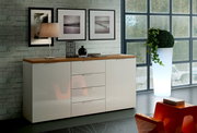 Modernes Sideboard mit glänzender weißer Front und Holzplatte, seitliche Perspektive, dekoriert mit Lampe und Vasen, vor einer Backsteinwand mit Bildern.