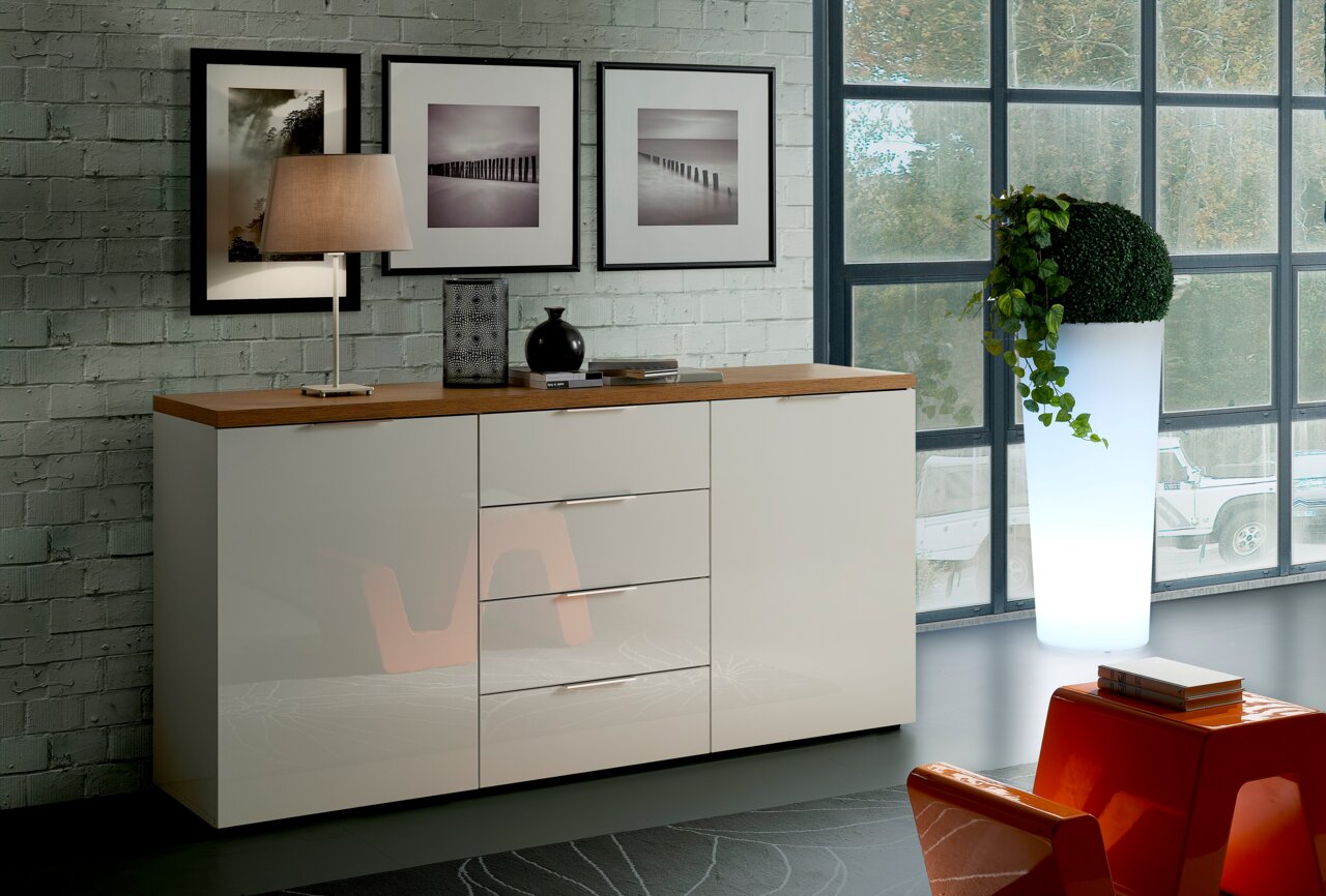 Modernes Sideboard mit glänzender weißer Front und Holzplatte, seitliche Perspektive, dekoriert mit Lampe und Vasen, vor einer Backsteinwand mit Bildern.