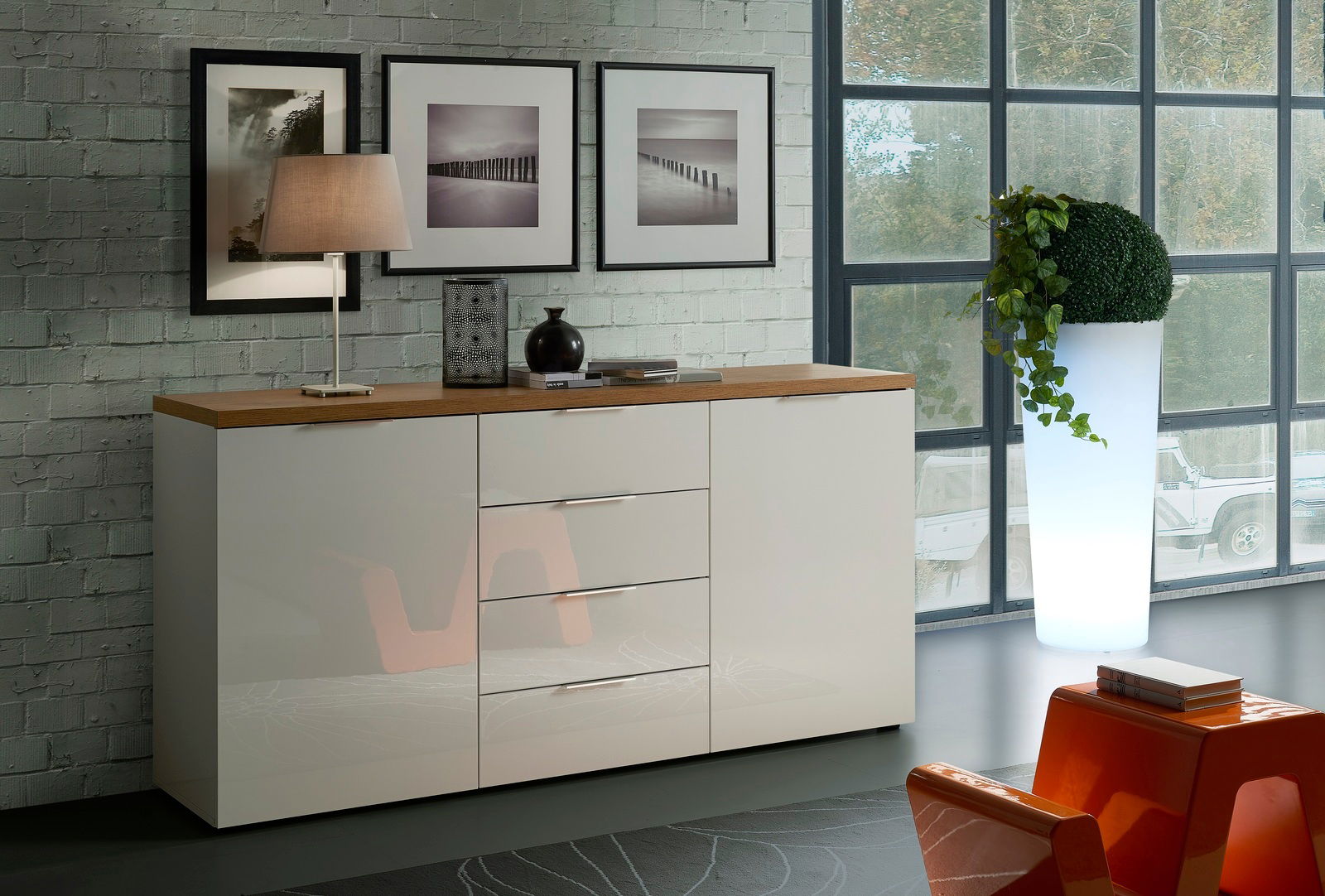 Modernes Sideboard mit glänzender weißer Front und Holzplatte, seitliche Perspektive, dekoriert mit Lampe und Vasen, vor einer Backsteinwand mit Bildern.