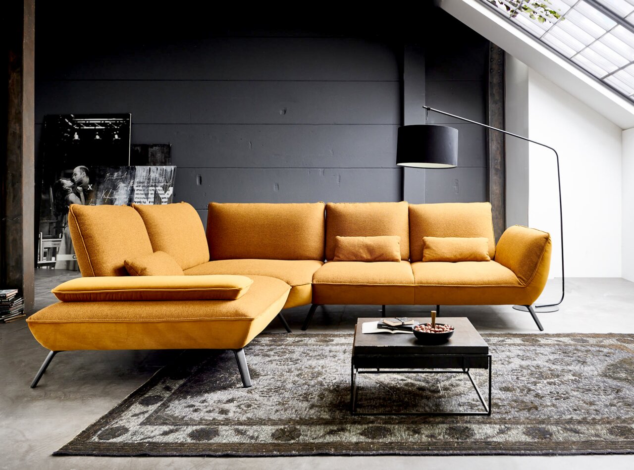 Moderano Ecksofa ALPHA Moderne Eckgarnitur in Senfgelb aus seitlicher Perspektive, platziert in einem stilvollen Wohnzimmer mit grauer Wand und großem Fenster.