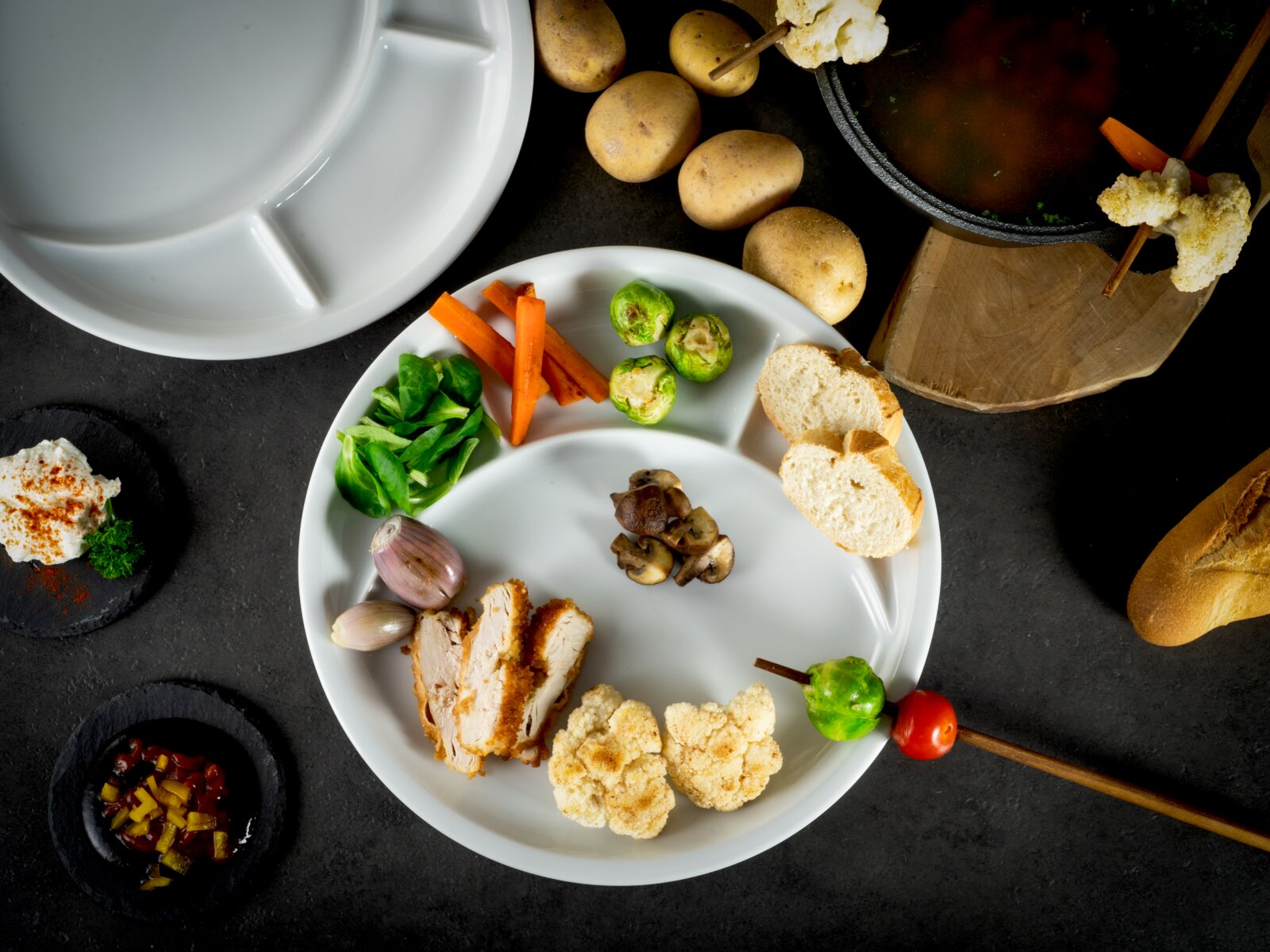 Draufsicht auf einen weißen, runden Menüteller mit mehreren Fächern, angerichtet mit Hähnchenschnitzelstreifen, Rosenkohl, Karottenstiften, Feldsalat, Champignons, Schalotten, Blumenkohl und Brotscheiben auf dunklem Untergrund; daneben Kartoffeln und Fond