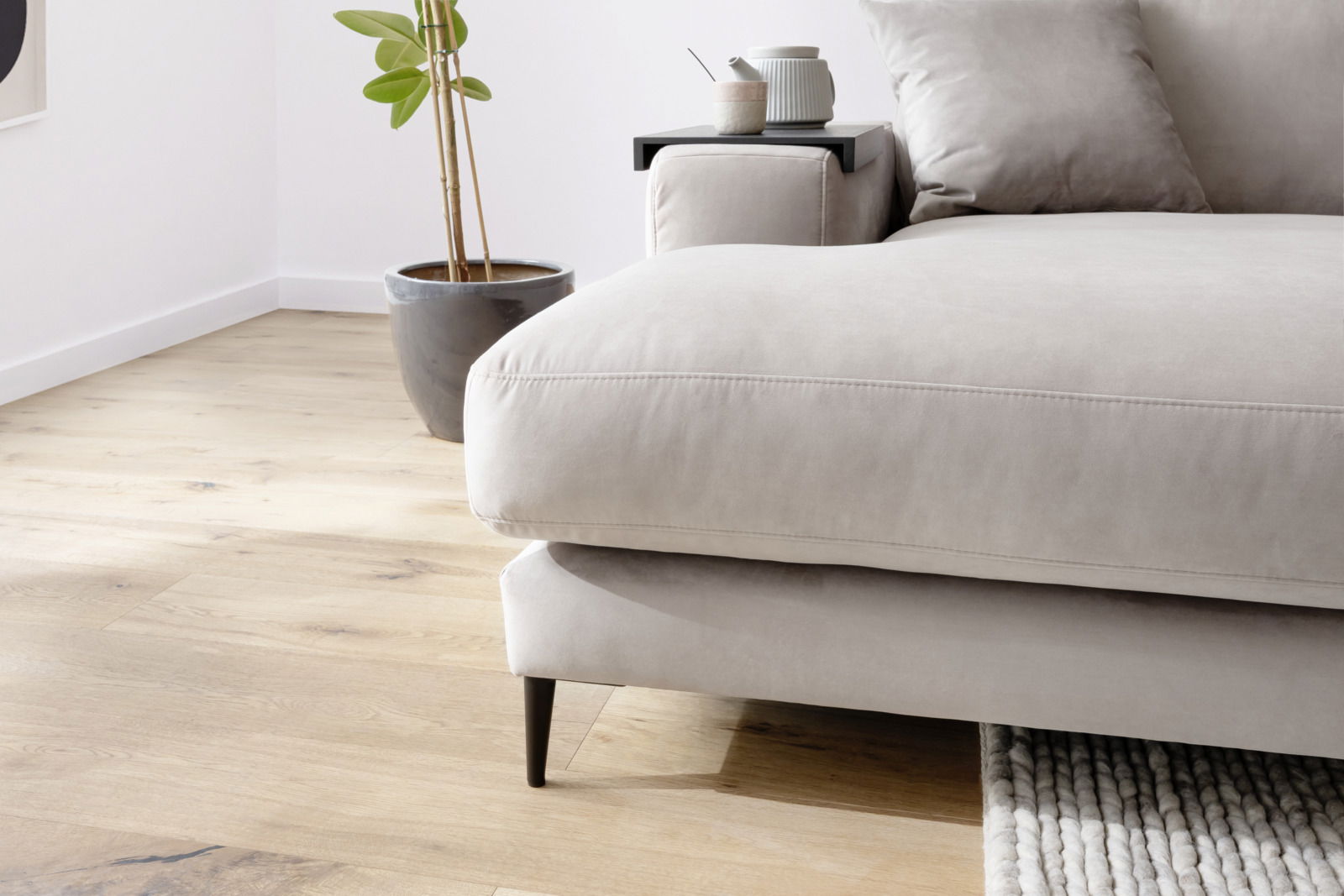 Nahaufnahme einer modernen Longchairkombination in hellem Beige, mit einem Kissen und einem Beistelltisch mit Teekanne und Tasse, auf hellem Holzboden. Perspektive von der Seite.