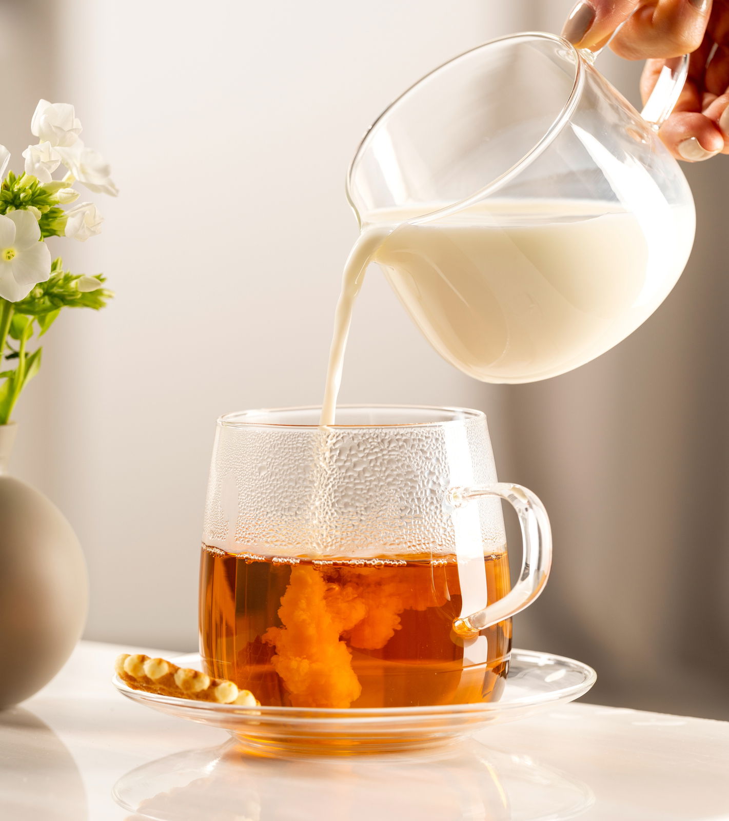 Seitliche Ansicht einer Untertasse mit einer Tasse Tee, in die Milch gegossen wird, neben einer kleinen Blume in einer Vase.