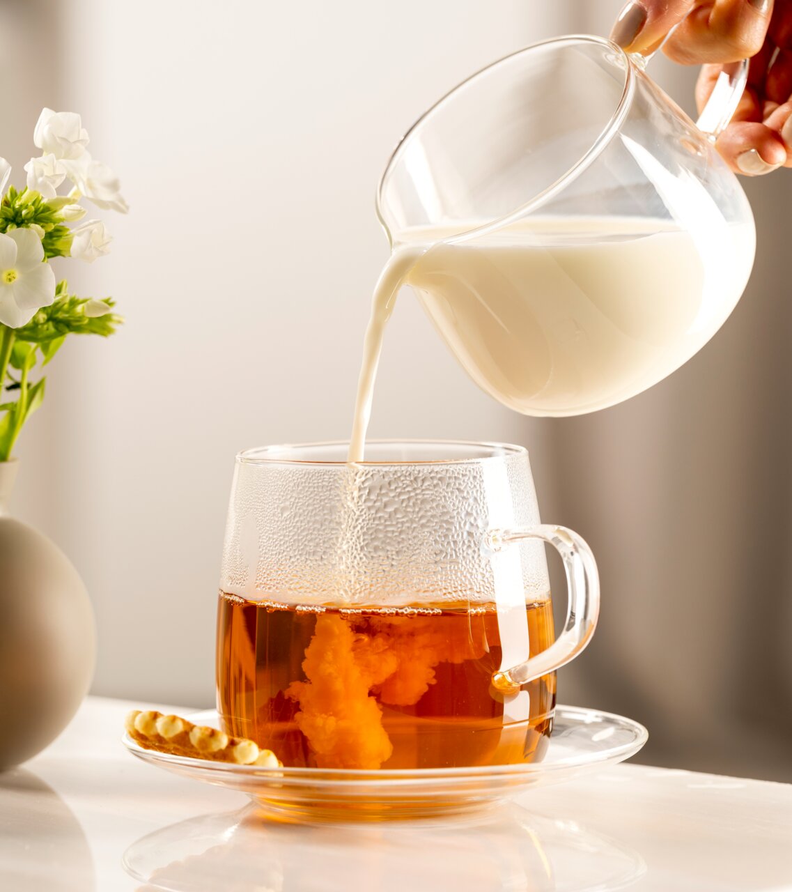 Seitliche Ansicht einer Untertasse mit einer Tasse Tee, in die Milch gegossen wird, neben einer kleinen Blume in einer Vase.